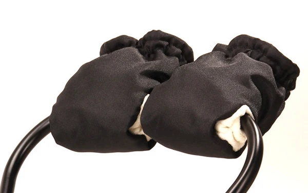 Close-up of black fleece-lined hand muffs attached to a stroller handle bar, lit with soft warm light against a clean white background, conveying a cozy, handcrafted feel.
